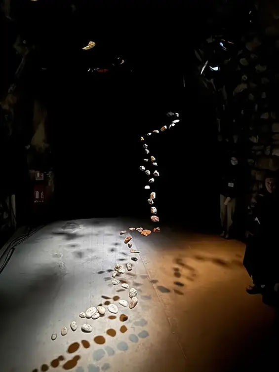 Clément Edouard & Pierce Warnecke – Artistes concepteurs Flux est une installation composée de roches suspendues mises en mouvement qui imagine l’histoire d’un cours d’eau disparu. Par des jeux de mouvements, de lumière et de son, la roche révèle ici la mémoire que l’eau lui a laissé́. Elle retranscrit sa fluidité́, sa vitesse et le parcours qu’elle a entamé́ du haut d’une montagne jusqu’à la mer. L’installation propose une compression spatiale et temporelle de la relation entre eau et roche, du granite des cimes au sable des deltas. Elle invite à une expérience sensible et imaginaire, soulignant l’interdépendance entre l’humain et le milieu ambiant. Les pierres ainsi que le tracé topographique et les matières sonores diffusées en quadriphonie ont été prélevés lors de différentes résidences aux Pays des Vans en Cévennes dans la rivière Le Chassezac (affluant principalement de l’Ardèche).
