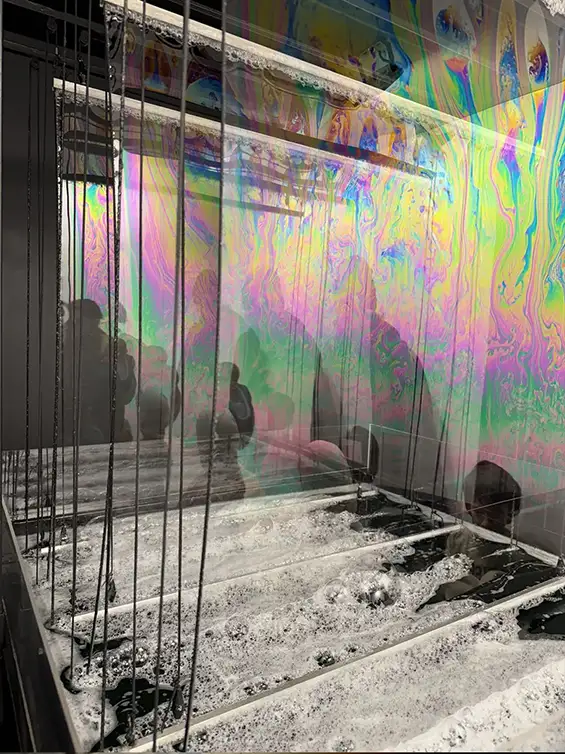Nicky Assmann – Artiste plasticienne : Solaris est une sculpture cinétique composée de plusieurs films de savon qui peuvent être soulevés manuellement. Il s’agit d’une expérience cinématographique qui explore non seulement le processus mental et l’activité physique de la vision mais également la création de films de savon dans l’espace. Nicky Assmann explore la perception visuelle à travers six écrans de films de savon placés les uns derrière les autres jouant ainsi avec la lumière et les reflets. Grâce à un éclairage précis, le mouvement interne du film de savon est révélé, montrant une chorégraphie turbulente de couleurs irisées et de mouvements fluides. La gravité prend lentement et inévitablement le contrôle de la membrane.